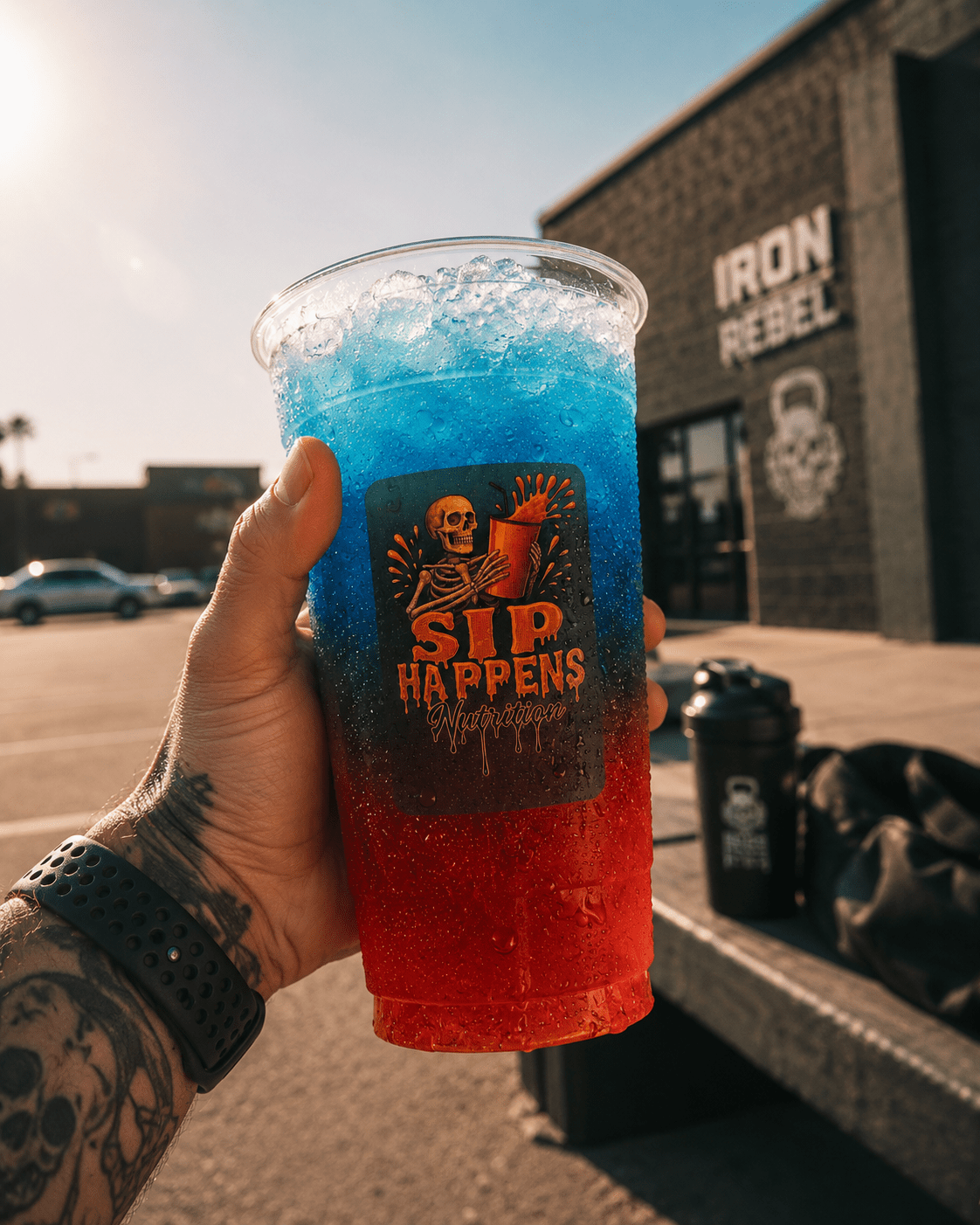 Tattooed hand holding a Sip Happens Nutrition iced drink with red-to-blue layers outside Iron Rebel gym