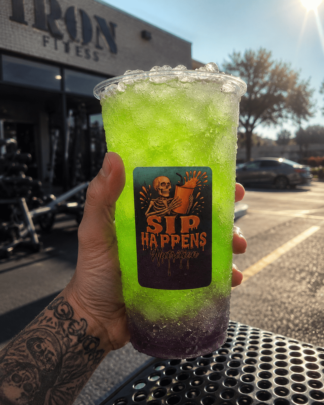 A tattooed hand holds a large iced drink with a bright green and purple gradient, branded with a Sip Happens Nutrition logo featuring a skeleton