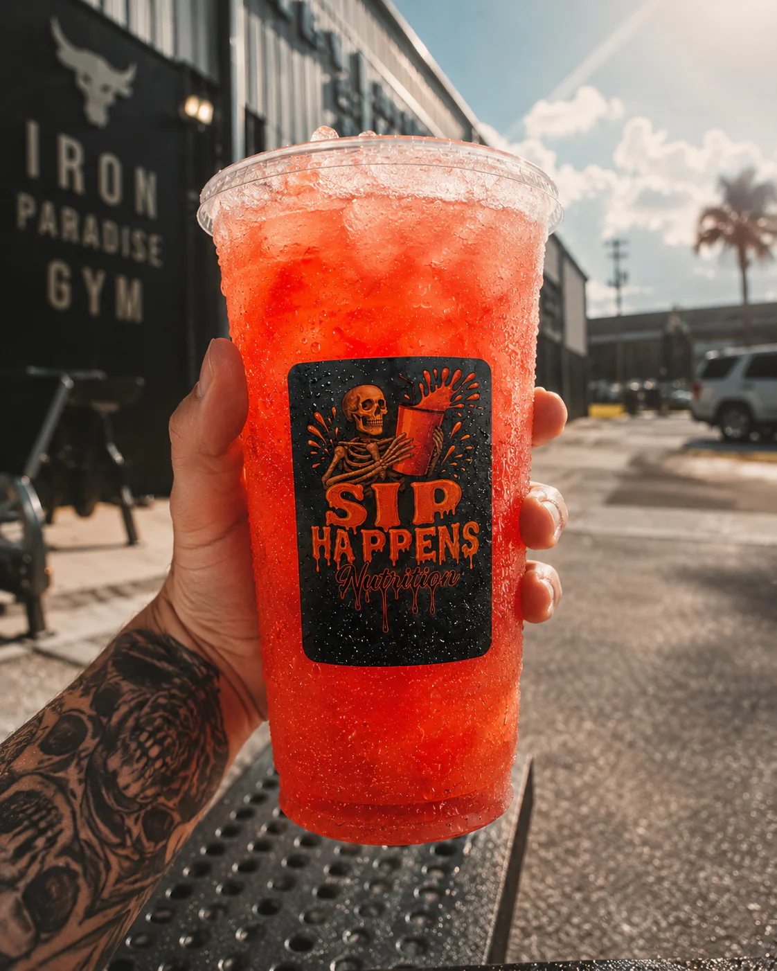 Tattooed hand holding a large iced red drink in a Sip Happens Nutrition cup outside Iron Paradise Gym