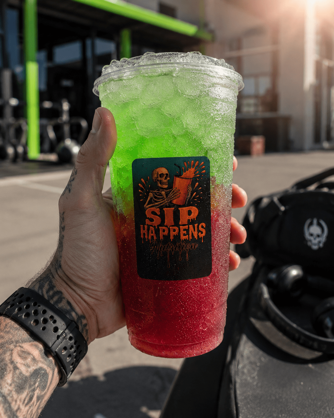 A tattooed hand holds a large iced drink with red-to-green gradient layers, branded with a Sip Happens Nutrition logo, outside a gym