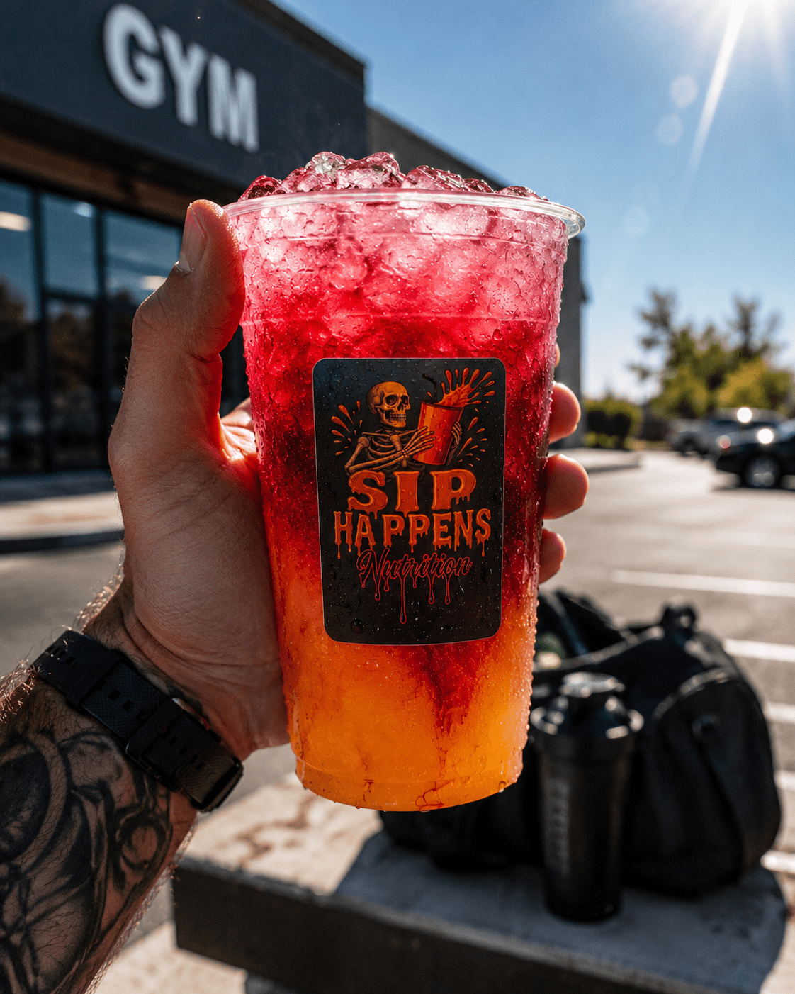 Tattooed hand holding a Sip Happens Nutrition iced drink with a red-to-orange gradient, outside a gym