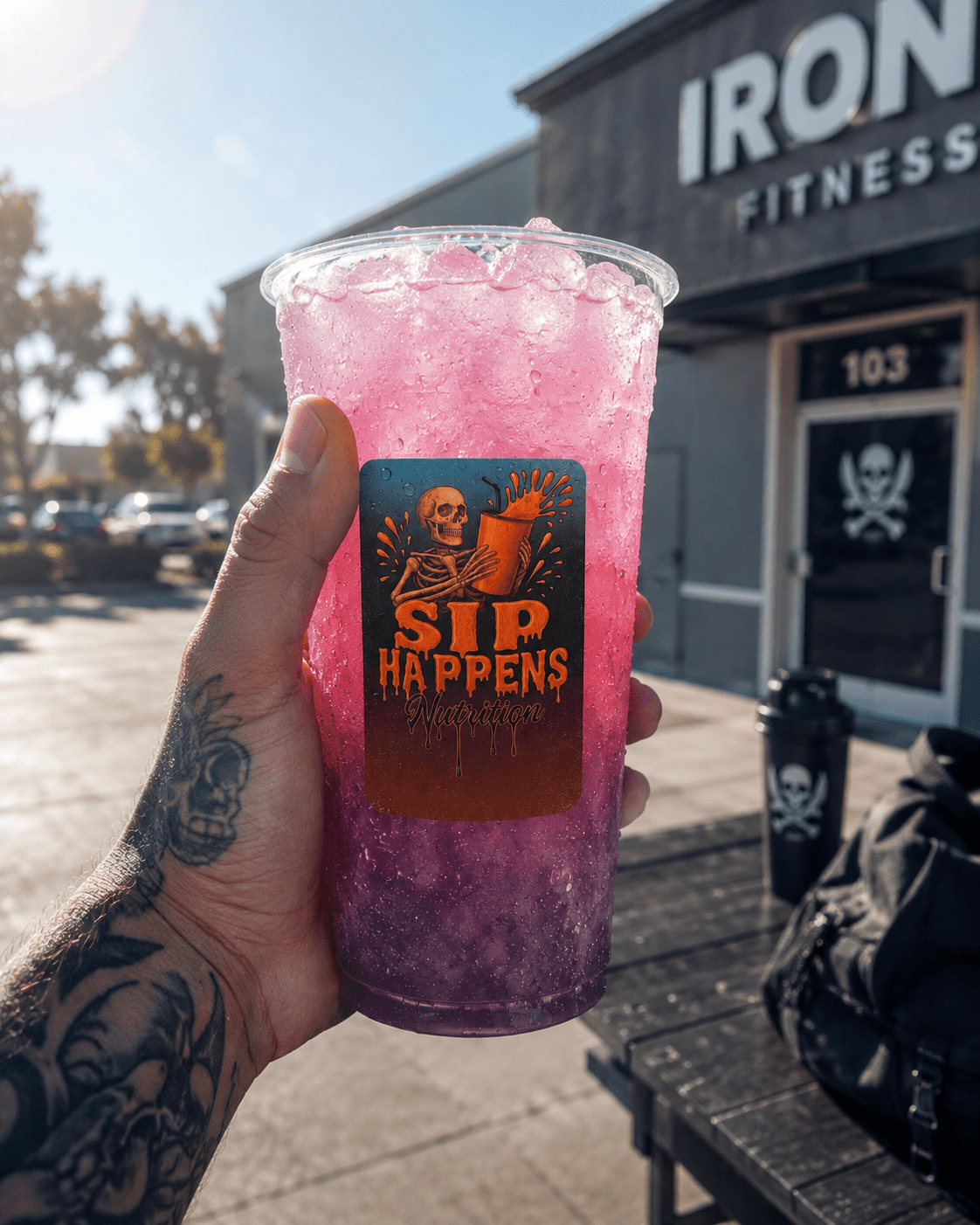 Tattooed hand holding a pink-to-purple iced drink labeled Sip Happens Nutrition outside Iron Fitness gym