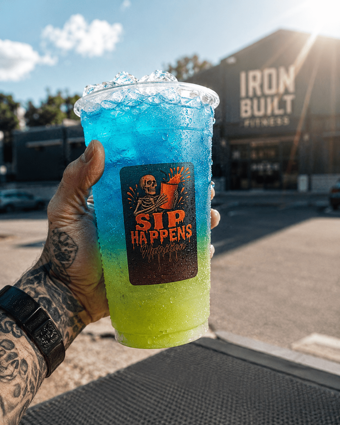 Tattooed hand holding a large Sip Happens Nutrition drink layered in blue and neon green, held up outside Iron Built Fitness