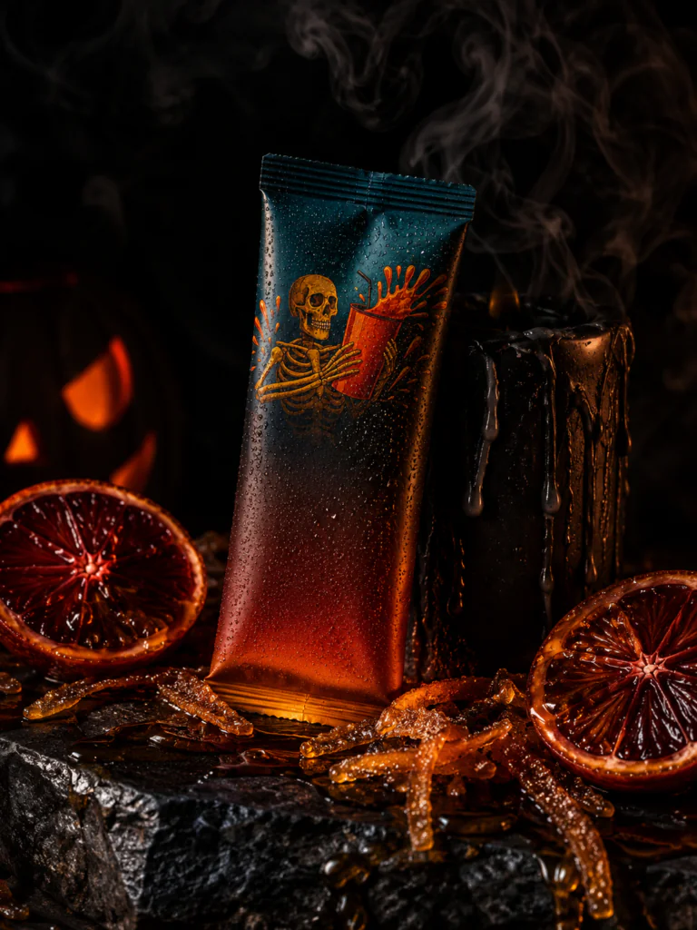 Dark-themed product pouch with skeleton illustration, surrounded by blood oranges, candied citrus peel, and a dripping black candle