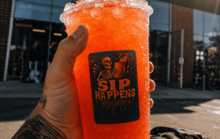 Tattooed hand holding a large orange iced drink from Sip Happens Nutrition in front of Ironcastle Fitness