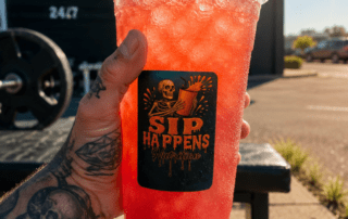 Tattooed hand holding a Sip Happens Nutrition iced red drink outside Iron Athletics 24/7 gym