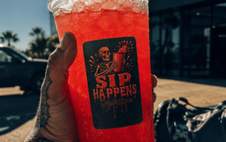 Tattooed hand holding a large red iced drink with a Sip Happens Nutrition logo outside Iron Will Fitness