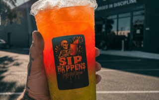 Tattooed hand holding a Sip Happens Nutrition drink with orange-to-green gradient outside Iron Legion Gym