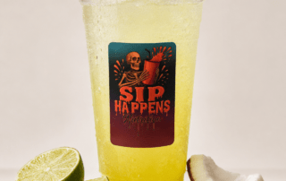 Sip Happens Nutrition iced drink with a yellow-green hue, surrounded by sliced limes, a lemon wedge, and coconut pieces