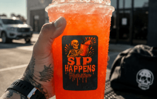 A tattooed hand holds a large orange iced drink branded Sip Happens Nutrition outside Iron Culture Gym
