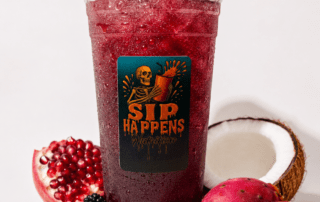 Sip Happens Nutrition iced drink in a large cup surrounded by pomegranate, blackberries, prickly pear, and coconut