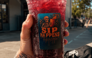 Tattooed hand holding a large iced red drink from Sip Happens Nutrition outside a fitness center